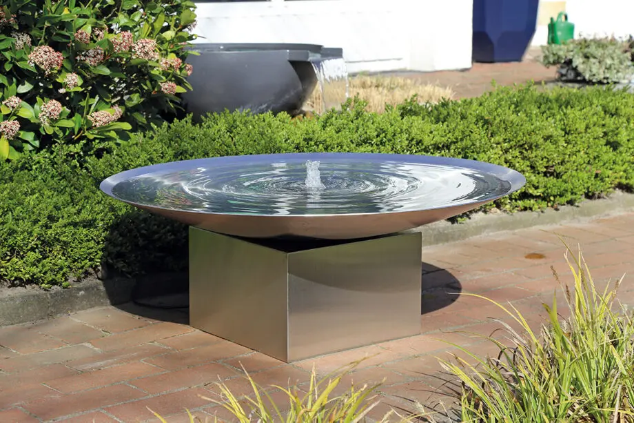Edelstahl-Wasserschale Esbjerg mit 100 cm Durchmesser auf einem Edelstahlsockel, sanfte Wasserfontäne, aufgestellt in einem modernen Garten mit Kies und Pflanzen.