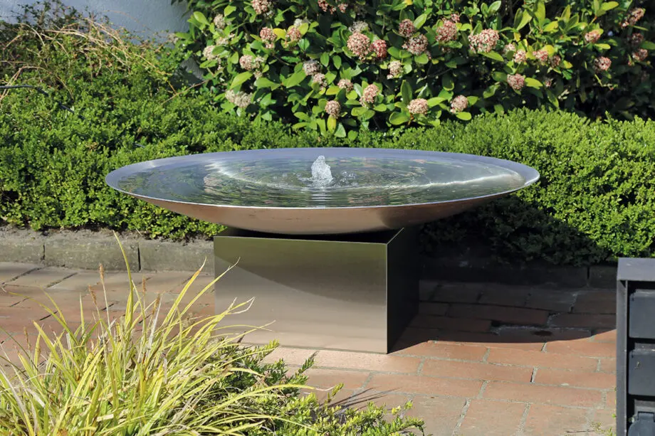 Edelstahl-Wasserschale Esbjerg mit 100 cm Durchmesser auf einem Edelstahlsockel, sanfte Wasserfontäne, aufgestellt in einem modernen Garten mit Kies und Pflanzen.