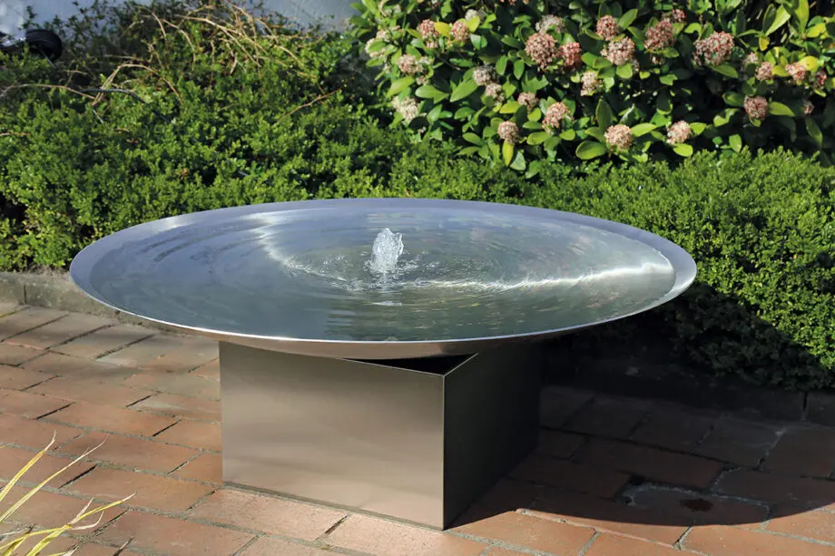 Edelstahl-Wasserschale Esbjerg mit 100 cm Durchmesser auf einem Edelstahlsockel, sanfte Wasserfontäne, aufgestellt in einem modernen Garten mit Kies und Pflanzen.