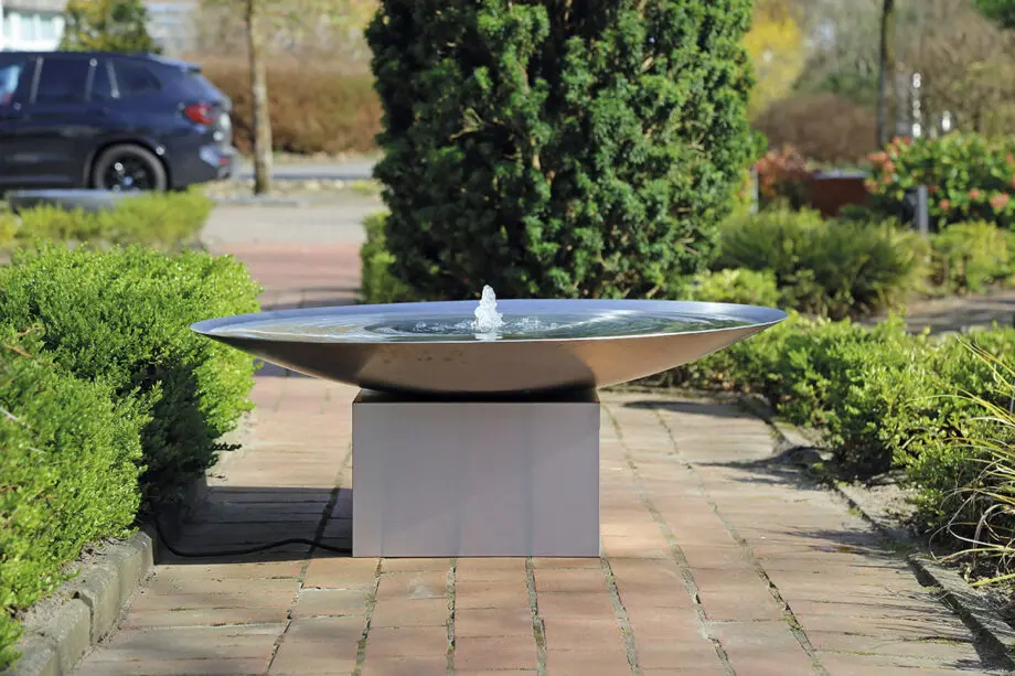 Edelstahl-Wasserschale Esbjerg mit 100 cm Durchmesser auf einem Edelstahlsockel, sanfte Wasserfontäne, aufgestellt in einem modernen Garten mit Kies und Pflanzen.