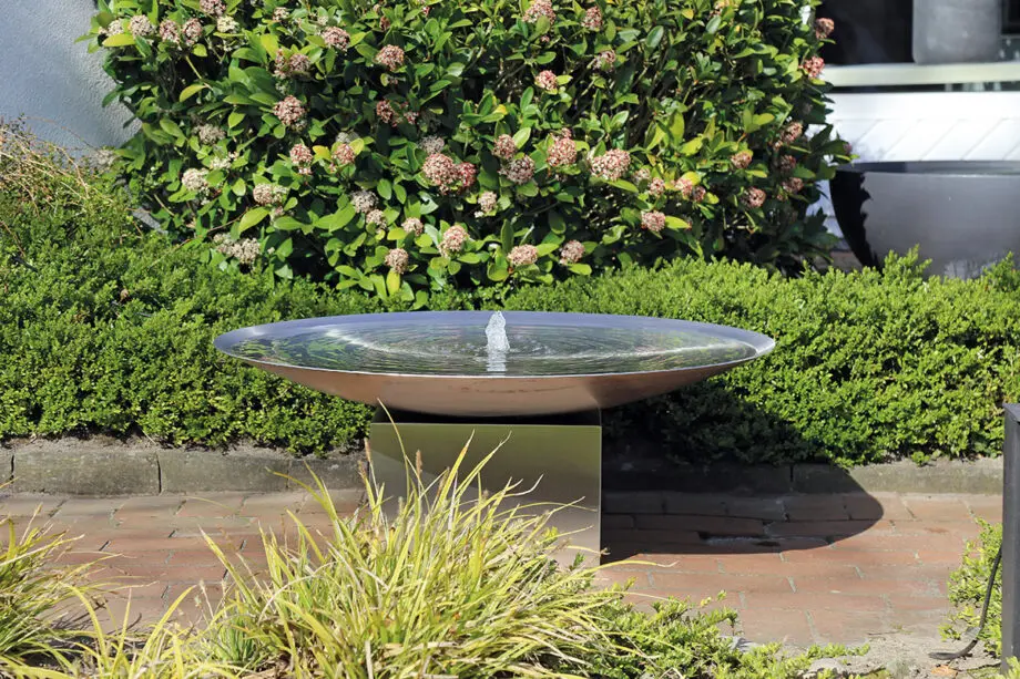 Edelstahl-Wasserschale Esbjerg mit 100 cm Durchmesser auf einem Edelstahlsockel, sanfte Wasserfontäne, aufgestellt in einem modernen Garten mit Kies und Pflanzen.