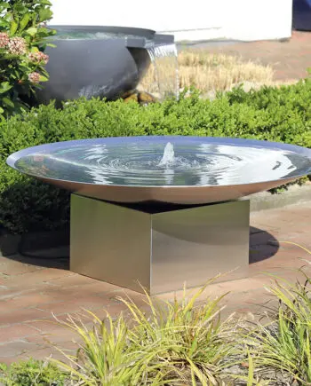 Edelstahl-Wasserschale Esbjerg mit 100 cm Durchmesser auf einem Edelstahlsockel, sanfte Wasserfontäne, aufgestellt in einem modernen Garten mit Kies und Pflanzen.