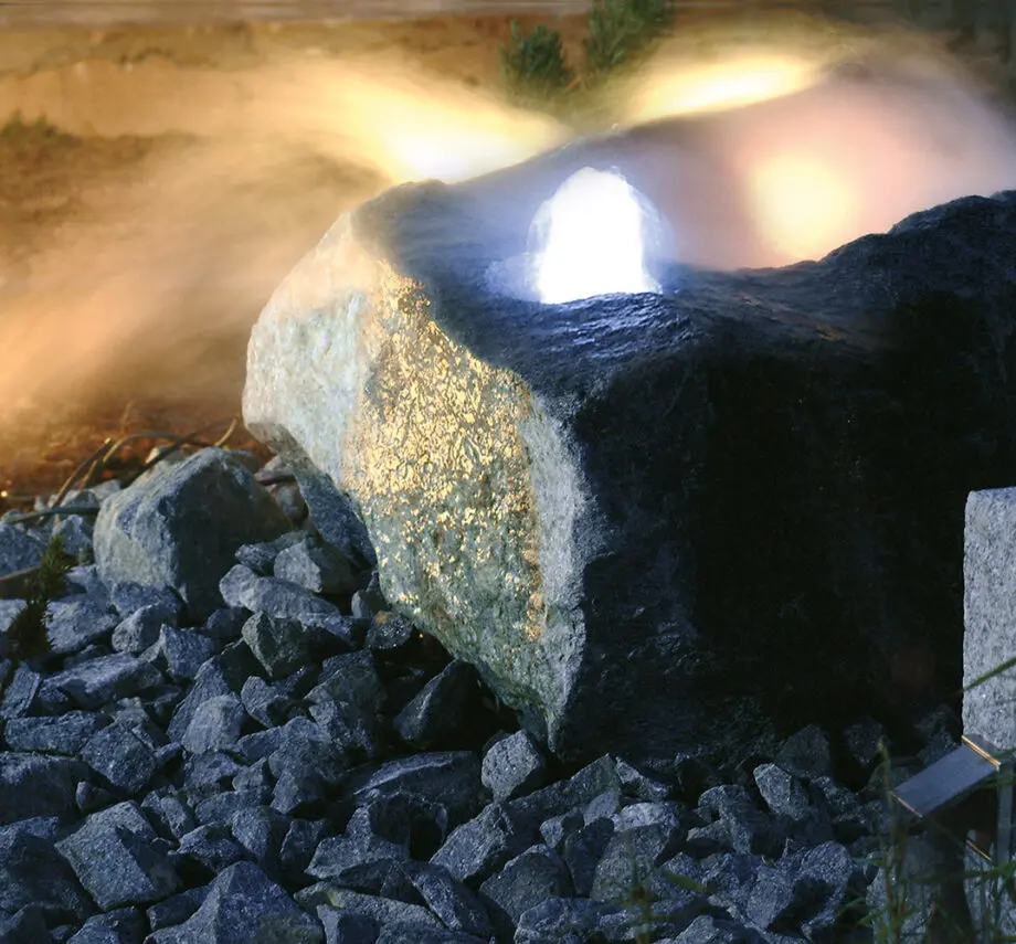 LED-Quellsteinleuchte 30 (warmweiß, 10 Lumen) beleuchtet einen Quellstein in einem Garten bei Dämmerung, mit sanftem Licht und fließendem Wasser.
