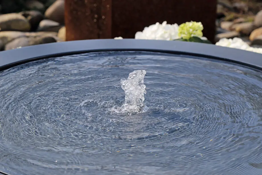 Wasserschale Lana in RAL-Farbe mit beleuchtetem Sprudelquell, Wasser bedeckt die Oberfläche, aufgestellt auf einer Terrasse bei Dämmerung, umgeben von modernen Pflanzen.