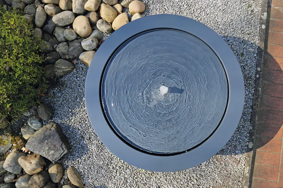 Wasserschale Lana in RAL-Farbe mit beleuchtetem Sprudelquell, Wasser bedeckt die Oberfläche, aufgestellt auf einer Terrasse bei Dämmerung, umgeben von modernen Pflanzen.