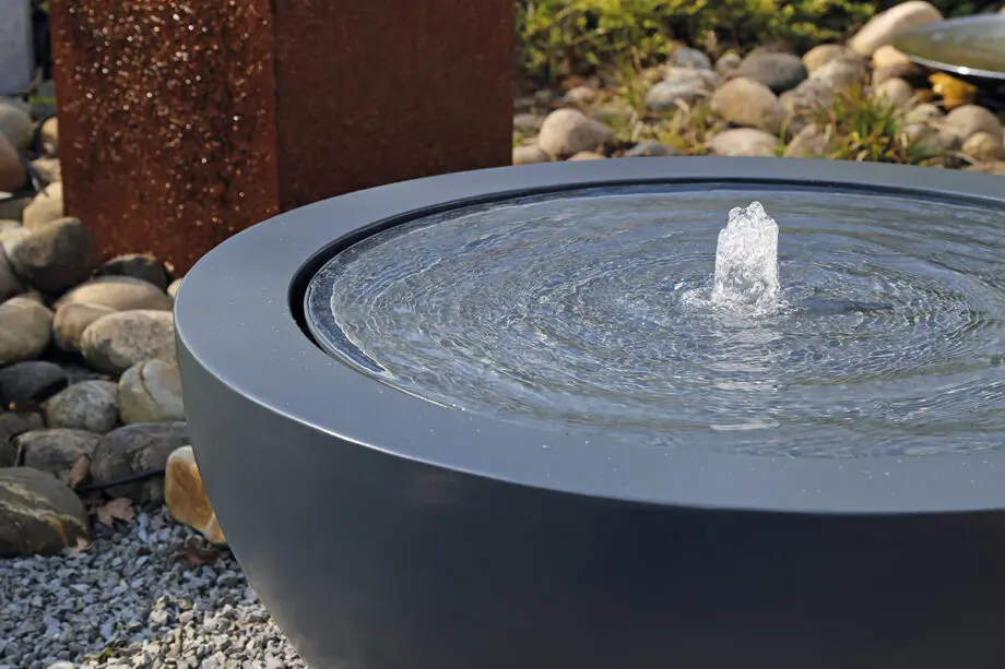 Wasserschale Lana in RAL-Farbe mit beleuchtetem Sprudelquell, Wasser bedeckt die Oberfläche, aufgestellt auf einer Terrasse bei Dämmerung, umgeben von modernen Pflanzen.