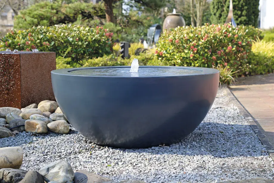 Wasserschale Lana in RAL-Farbe mit beleuchtetem Sprudelquell, Wasser bedeckt die Oberfläche, aufgestellt auf einer Terrasse bei Dämmerung, umgeben von modernen Pflanzen.