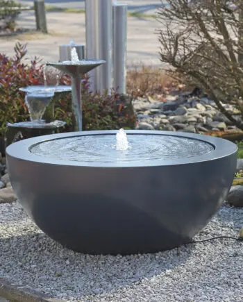 Wasserschale Lana in RAL-Farbe mit beleuchtetem Sprudelquell, Wasser bedeckt die Oberfläche, aufgestellt auf einer Terrasse bei Dämmerung, umgeben von modernen Pflanzen.