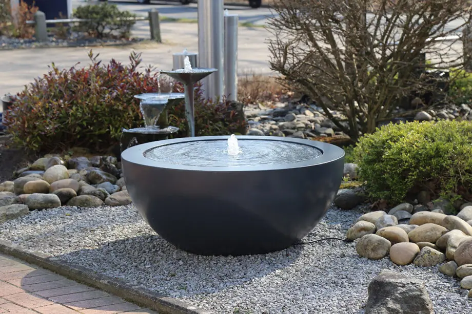 Wasserschale Lana in RAL-Farbe mit beleuchtetem Sprudelquell, Wasser bedeckt die Oberfläche, aufgestellt auf einer Terrasse bei Dämmerung, umgeben von modernen Pflanzen.