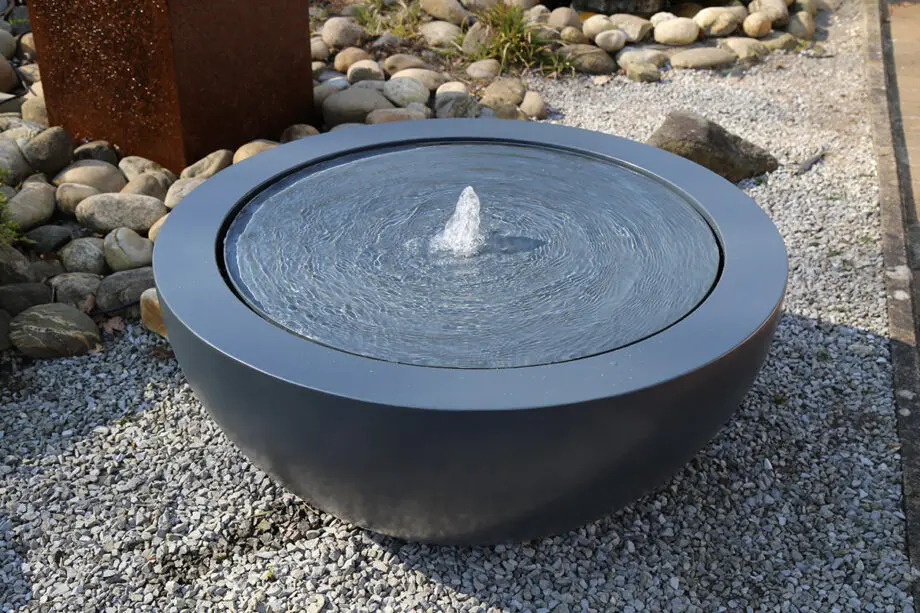 Wasserschale Lana in RAL-Farbe mit beleuchtetem Sprudelquell, Wasser bedeckt die Oberfläche, aufgestellt auf einer Terrasse bei Dämmerung, umgeben von modernen Pflanzen.