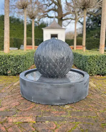 Zinkbrunnen Artischocke Modell 3551 mit kunstvoller Kugelstruktur (Ø 70 cm), aufgestellt in einem klassischen Garten mit runden Kieswegen und grünen Sträuchern.