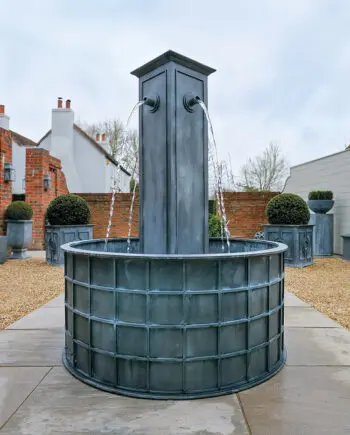 Zinkbrunnen Zürich (130 x 130 x 180 cm) mit hoher Mittelsäule und vier Wasserauslässen, aufgestellt in einem klassischen Garten mit gepflegtem Rasen und Steinwegen.