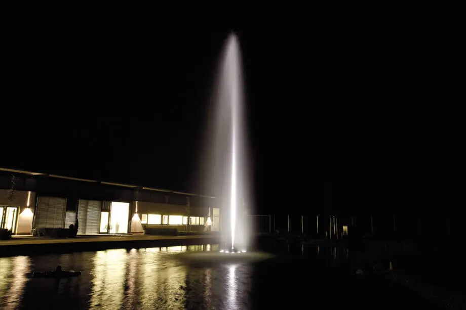 High Jet 30 auf einer Schwimmfontäne Profi in einem großen Teich, mit einem präzisen vertikalen Wasserstrahl aus Edelstahldüse, umgeben von Schilf und Bäumen.