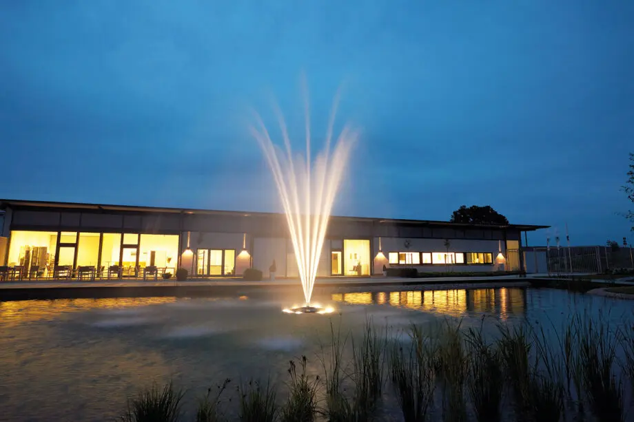 Oase Grand Vulkan 30 auf einer Schwimmfontäne Profi in einem Teich, mit einem kelchförmigen Wasserbild aus kreisförmigen Einzelstrahlen, umgeben von grünen Uferpflanzen.