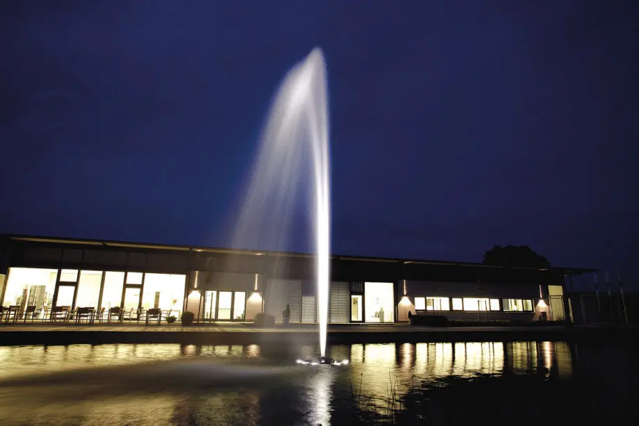 Oase Geyser Jet 30 auf einer Schwimmfontäne Profi in einem Teich, mit einem 5 m hohen, breiten Wasserstrahl aus Edelstahldüse, umgeben von grünen Uferpflanzen.