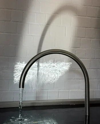 Modern geschwungener Wasserauslauf Kyoto aus gebürstetem Edelstahl, montiert über einem dunklen Wasserbecken mit Lichtreflexen an einer weißen Ziegelwand. Eine stimmungsvolle Lichtquelle wirft den Schatten des Bogens und Wellenreflexionen auf die Mauer.